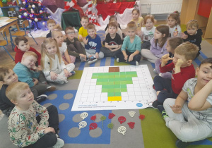 Dzieci siedzą na dywanie przed matą do kodowania, na której znajduje się ułożona choinka z zielonych kartoników. Obok maty leży kilka ozdób choinkowych.