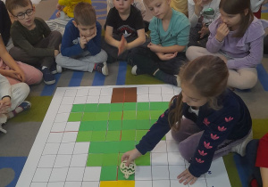 Dzieci siedzą na dywanie obok maty do kodowania, na której znajduje się ułożona choinka. Amanda układa na choince bombkę zgodnie z podaną instrukcją.