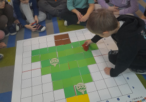 Na macie do kodowania ułożony jest obrazek choinki. Hubert przykucnął obok maty i układa na choince dzwoneczek zgodnie z podanym warunkiem. Dzieci obserwują działanie kolegi.