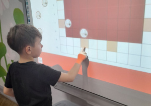 Staś stoi przed tablicą multimedialną, na której wyświetlony jest stół wigilijny. Chłopiec nakrywa stół przesuwając obrazki w określone miejsca.