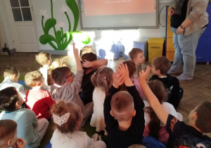 Dzieci siedzą na dywanie przed tablicą multimedialną, na której jest wyświetlony slajd prezentacji. Obok tablicy stoi Pani psycholog, która zadaje pytanie dzieciom. Przedszkolaki zgłaszają się do odpowiedzi poprzez podnoszoną w górę rękę.