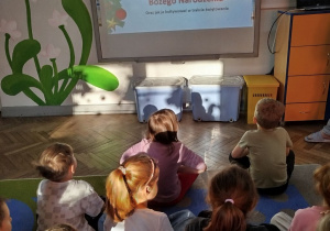 Dzieci siedzą na dywanie przed tablicą multimedialną ,na której wyświetlony jest slajd prezentacji.