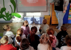 Dzieci siedzą na dywanie przed tablicą multimedialną, na której wyświetlony jest slajd prezentacji mówiący o tradycji. Obok tablicy stoi pani psycholog, która opowiada o tym, jakie mamy tradycje świąteczne.