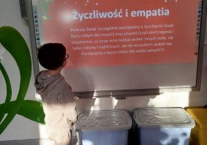 Mikołaj stoi przed tablicą multimedialną i czyta głośno slajd na temat wartości - życzliwości i empatii.