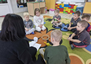 Dzieci razem z panią Karoliną siedzą na dywanie. Przed nimi leża tamburyn, marakasy i zgniecione kartki papieru.