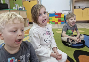 Karol, Weronika i Olek siedzą na dywanie. Dzieci ćwiczą wyciągając język na boki.