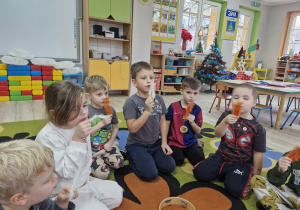 Dzieci siedząc na dywanie dmuchają na trzymane w dłoniach kolorowe piórka.