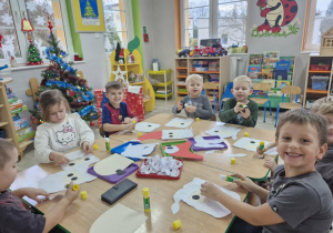 Grupa dzieci siedzi przy stoliczku, na którym leżą elementy wycięte z papieru kolorowego oraz kleje.