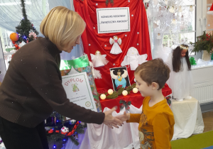 Pani dyrektor wręcza Bartkowi dyplom i nagrodę. W tle wystawa prac konkursowych oraz tablica z napisem - Konkurs rodzinny "Świąteczny Aniołek".