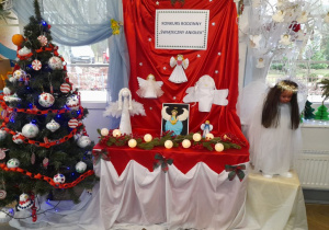 Wystawa prac konkursowych - na tablicy przykrytej czerwonym materiałem przypięty jest napis Konkurs rodzinny "Świąteczny Aniołek", trzy Aniołki oraz gwiazdki. Na szafce pod tablicą stoją cztery Aniołki wykonane przez dzieci i rodziców. Z lewej strony stoi choinka.