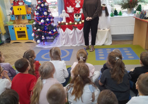 Dzieci siedzą na dywanie, a przed nimi stoi Pani dyrektor. W tle wystawa prac konkursowych, choinka, kącik domowy, okno z dekoracją świąteczną.