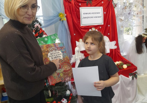 Nadia trzyma dyplom, a Pani dyrektor pokazuje nagrodę w postaci książki - gry dla poszukiwaczy przygód pt. "Miasto Tańczącego Karpia".