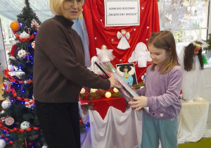 Weronika odbiera nagrodę od Pani dyrektor za udział w konkursie. W tle wystawa prac konkursowych, tablica z napisem Konkurs rodzinny "Świąteczny Aniołek", choinka.
