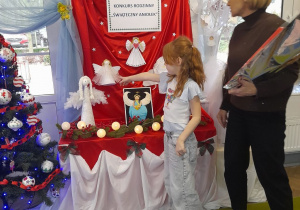 Emma wskazuje przypiętego na tablicy swojego Aniołka, a obok stoi Pani dyrektor trzymając nagrodę dla dziewczynki.