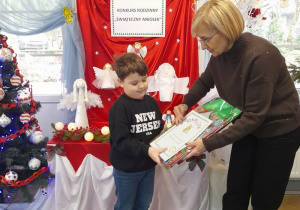 Pani dyrektor wręcza Julkowi dyplom i nagrodę w świątecznej torebce. W tle tablica z napisem - Konkurs rodzinny "Świąteczny Aniołek" oraz wystawa prac konkursowych.