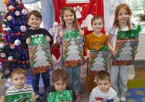 Dzieci pozują do wspólnego zdjęcia z nagrodami zapakowanymi w świąteczne torebki. W tle wystawa prac konkursowych, tablica z napisem - Konkurs rodzinny "Świąteczny Aniołek", choinka.