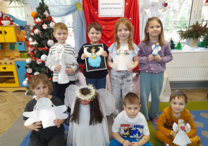 Dzieci prezentują wykonane Aniołki. W tle tablica z napisem - Konkurs rodzinny "Świąteczny Aniołek", choinka, okno z dekoracją świąteczną.