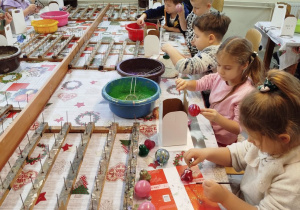 Dzieci siedzą przy wspólnym stole podczas warsztatów zdobienia bombek. Przedszkolaki malują bombki klejem i obsypują brokatem.