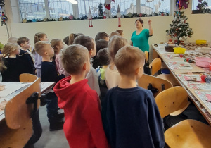 Dzieci podczas zajęć warsztatowych stoją przed Panią, która omawia sposób dekorowania bombek.