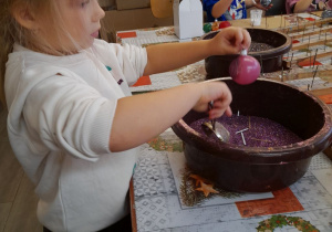 Tosia obsypuje swoją bombkę brokatem. W tle Mikołaj i Emma malują bombki klejem.