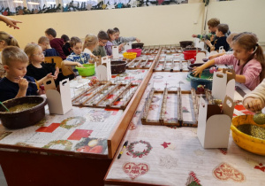 Dzieci siedzą przy stole podczas ozdabiania bombek. Na środku leżą suszarki do bombek, a przed dziećmi pojemniki na bombki.