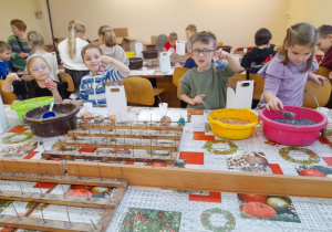 Dzieci siedzą przy stołach i ozdabiają swoje bombki. Bartek i Mikołaj podnoszą bombki do góry. Przed dziećmi leży suszarka do bombek.