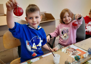 Mikołaj i Emma siedzą przy stole i podnoszą do góry ozdobione przez siebie bombki.