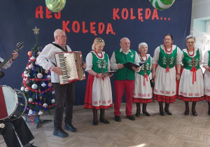 Na scenie stoi Ludowy Zespół Wokalno-Instrumentalny "Leszczynianki". W tle napis "Hej Kolęda, Kolęda..." przypięty na granatowym materiale. Obok stoi ubrana choinka.