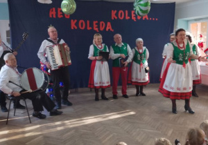 Pani w stroju ludowym stoi na środku sceny i opowiada dzieciom o zespole. W tle pozostali członkowie zespołu.