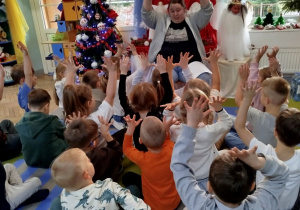 Pani psycholog siedzi przed dekoracją świąteczną, a przed nią na dywanie siedzą dzieci. Wszystkie dzieci podnoszą do góry ręce z otwartymi dłońmi podczas rozmowy o emocjach w wybranych sytuacjach świątecznych.