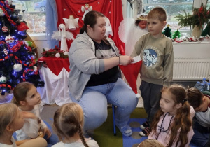 Mikołaj stoi przed Panią psycholog i losuje z czapki świątecznej hasło do odtworzenia podczas gry w kalambury. Przed nimi dzieci siedzą na dywanie.