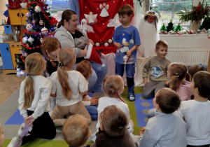 Staś stoi obok Pani psycholog i pokazuje wylosowaną czynność podczas gry w kalambury. Dzieci siedzą przed nimi na dywanie i próbują odgadnąć hasło.