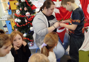 Marcel stoi przed Panią psycholog i losuje hasło do odegrania podczas gry w kalambury. Przed nimi dzieci siedzą na dywanie.