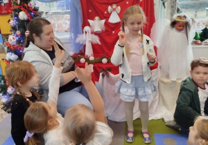 Nikola stoi obok Pani psycholog i pokazuje dzieciom wylosowaną czynność. Przed nimi siedzą dzieci na dywanie z podniesionymi rękami, chcąc zgadnąć hasło.