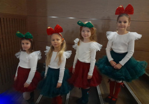 Cztery dziewczynki w oczekiwaniu na występ stoją jedno za drugim na schodach w sali widowiskowej Kutnowskiego Domu Kultury. Dziewczynki ubrane są w czerwone i zielone spódniczki, kremowe bluzeczki, czerwone i zielone podkolanówki, a na głowach mają opaski z kokardami.