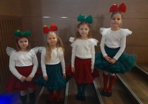 Cztery dziewczynki w oczekiwaniu na występ stoją obok siebie na schodach w sali widowiskowej Kutnowskiego Domu Kultury. Dziewczynki ubrane są w czerwone i zielone spódniczki, kremowe bluzeczki, czerwone i zielone podkolanówki, a na głowach mają opaski z kokardami.
