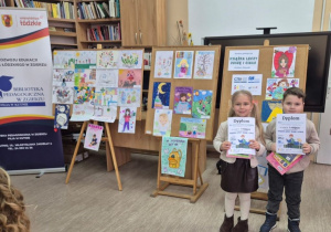 Tosia i Julek stoją obok siebie po ogłoszeniu wyników konkursu w Bibliotece Pedagogicznej. Dzieci trzymają przed sobą dyplomy i nagrody książkowe. W tle wystawa prac konkursowych.