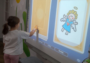 Nadia stoi przy tablicy multimedialnej i odkrywa kartę do gry.