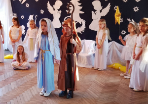 Marysia w roli Maryi i Antek w roli Józefa stoją na środku sceny i odtwarzają dalszą część przedstawienia. W tle fragment dekoracji jasełkowej, dziewczynki w roli Aniołków i Gwiazdek.