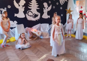 Na środku sceny stoi Tosia w roli Aniołka. W tle fragment dekoracji jasełkowej, dziewczynki w roli Aniołków i Gwiazdek, Mikołaj w roli Gospodarza.