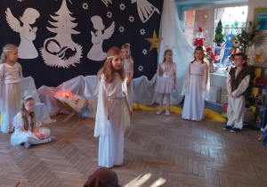 Emma stoi na środku sceny i odtwarza rolę Aniołka. W tle fragment dekoracji jasełkowej, dziewczynki w roli Aniołków i Gwiazdek, Mikołaj w roli Gospodarza. Z prawej strony siedzą trzej Królowie.