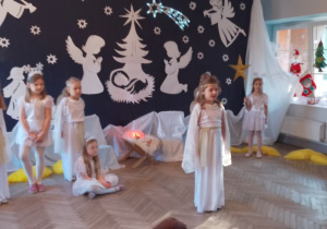 Nadia w roli Aniołka stoi na środku sceny. W tle fragment dekoracji jasełkowej, dziewczynki w roli Aniołków i Gwiazdek. Z prawej strony Mikołaj w roli Gospodarza.