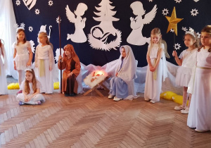 Przy żłóbku siedzi Marysia przebrana za Maryję i Antek jako Józef. Po bokach stoją dziewczynki w roli w roli Aniołków i Gwiazdek. W tle dekoracja.