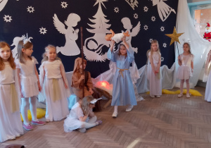 Marysia w roli Maryi idzie z lalką - Jezuskiem podnosząc go do góry. Dziewczynka odtwarza fragment przedstawienie. Obok stoją dziewczynki przebrane za Aniołki i Gwiazdki, a przy żłóbku siedzi Antek w roli Józefa.