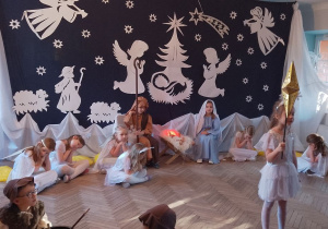 Amanda w roli Gwiazdki stoi na środku sceny i prezentuje fragment inscenizacji. Z tyłu śpią Aniołki i Gwiazdki, a obok żłóbka siedzi Maryja i Józef. W tle dekoracja - na granatowym tle przypięte są postacie aniołków, baranków, pastuszka, żłóbek, gwiazdki. Na górze świeci gwiazda betlejemska.