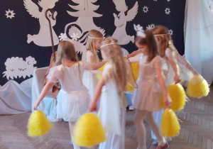 Dziewczynki w roli Gwiazdek i Aniołków tańczą z pomponami przy muzyce. W tle fragment dekoracji.