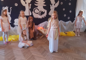 Nikola w roli Aniołka prezentuje fragment inscenizacji na środku sceny. W tle dzieci w roli Aniołków, Gwiazdek, Józefa oraz dekoracja jasełkowa.