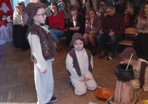 Mikołaj w roli Pasterza stoi przy ognisku prezentując swoją rolę. W tle rodzice siedzą na krzesłach.