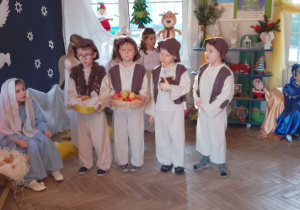 Czterech chłopców w roli Pasterzy stoi obok żłóbka. Chłopcy trzymają dary dla Jezusa: bochen chleba, koszyk z jabłkami, garnuszek ze śmietaną. Obok żłóbka siedzi Maryja, a w tle trzech królów.