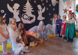 Królowie stoją obok żłóbka z darami w rękach. Przy żłobku siedzą Maryja i Józef, a obok stoją Aniołki i Gwiazdki. W tle dekoracja jasełkowa.